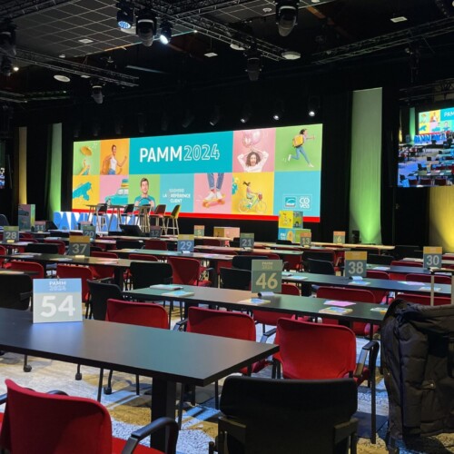 La performance audiovisuelle réalisée par TEDELEC Event à l'occasion du PAMM 2024 a mis en lumière l'excellence technique avec des éclairages dynamiques, un affichage LED de haute qualité et une configuration immersive adaptée à un événement d'envergure. Vue d'une salle de conférence avec des chaises rouges et des tables noires, prêtes pour l'événement PAMM 2024. Une scène avec un grand écran LED multicolore affichant le logo de l'événement est visible, avec plusieurs projecteurs éclairant la scène et des écrans supplémentaires affichant la scène en temps réel.
