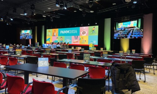 La performance audiovisuelle réalisée par TEDELEC Event à l'occasion du PAMM 2024 a mis en lumière l'excellence technique avec des éclairages dynamiques, un affichage LED de haute qualité et une configuration immersive adaptée à un événement d'envergure. Vue d'une salle de conférence avec des chaises rouges et des tables noires, prêtes pour l'événement PAMM 2024. Une scène avec un grand écran LED multicolore affichant le logo de l'événement est visible, avec plusieurs projecteurs éclairant la scène et des écrans supplémentaires affichant la scène en temps réel.