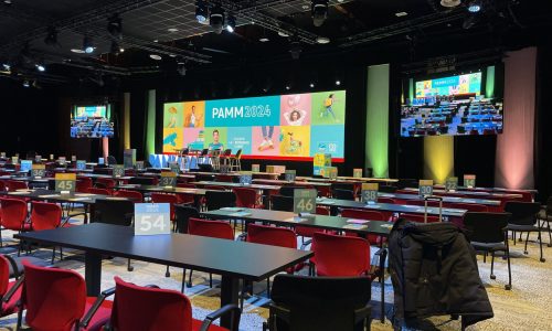 La performance audiovisuelle réalisée par TEDELEC Event à l'occasion du PAMM 2024 a mis en lumière l'excellence technique avec des éclairages dynamiques, un affichage LED de haute qualité et une configuration immersive adaptée à un événement d'envergure. Vue d'une salle de conférence avec des chaises rouges et des tables noires, prêtes pour l'événement PAMM 2024. Une scène avec un grand écran LED multicolore affichant le logo de l'événement est visible, avec plusieurs projecteurs éclairant la scène et des écrans supplémentaires affichant la scène en temps réel.